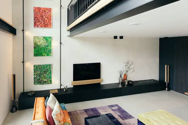 Strakke, lichte woonkamer met een naadloze lichte wand als achtergrond. Een zwart lowboard integreert TV en opbergruimte, kleurrijke kunstwerken en goudkleurige accenten voegen warme details toe.