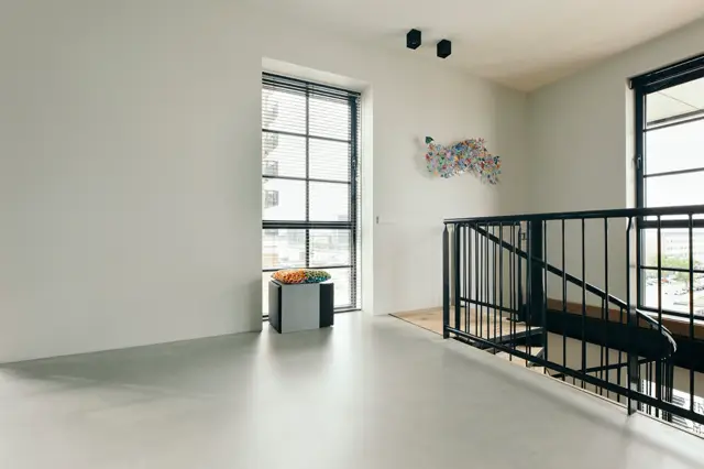 Ruim, lichtgevuld loft-achtig interieur met een doorlopende lichte naadloze vloer. Strakke witte wanden, grote ramen met zwarte kozijnen, minimalistische inrichting en een zwarte trap creëren een modern, opgeruimd geheel.