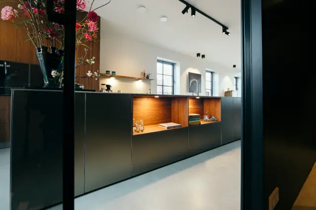 Een strak, licht interieur met naadloze vloer in natuurlijke tint, zwarte meubels met houten details en ingebouwde verlichting. De ruimte straalt rust en verfijning uit met natuurlijk licht en geordende styling.