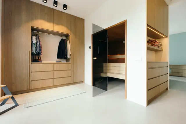 Ruime inloopkast met eiken fronten, grijze kastdeuren en open kledingrek. Naadloze witte vloer creëert een airy, minimalistische uitstraling. Warm licht en strakke architectuur.