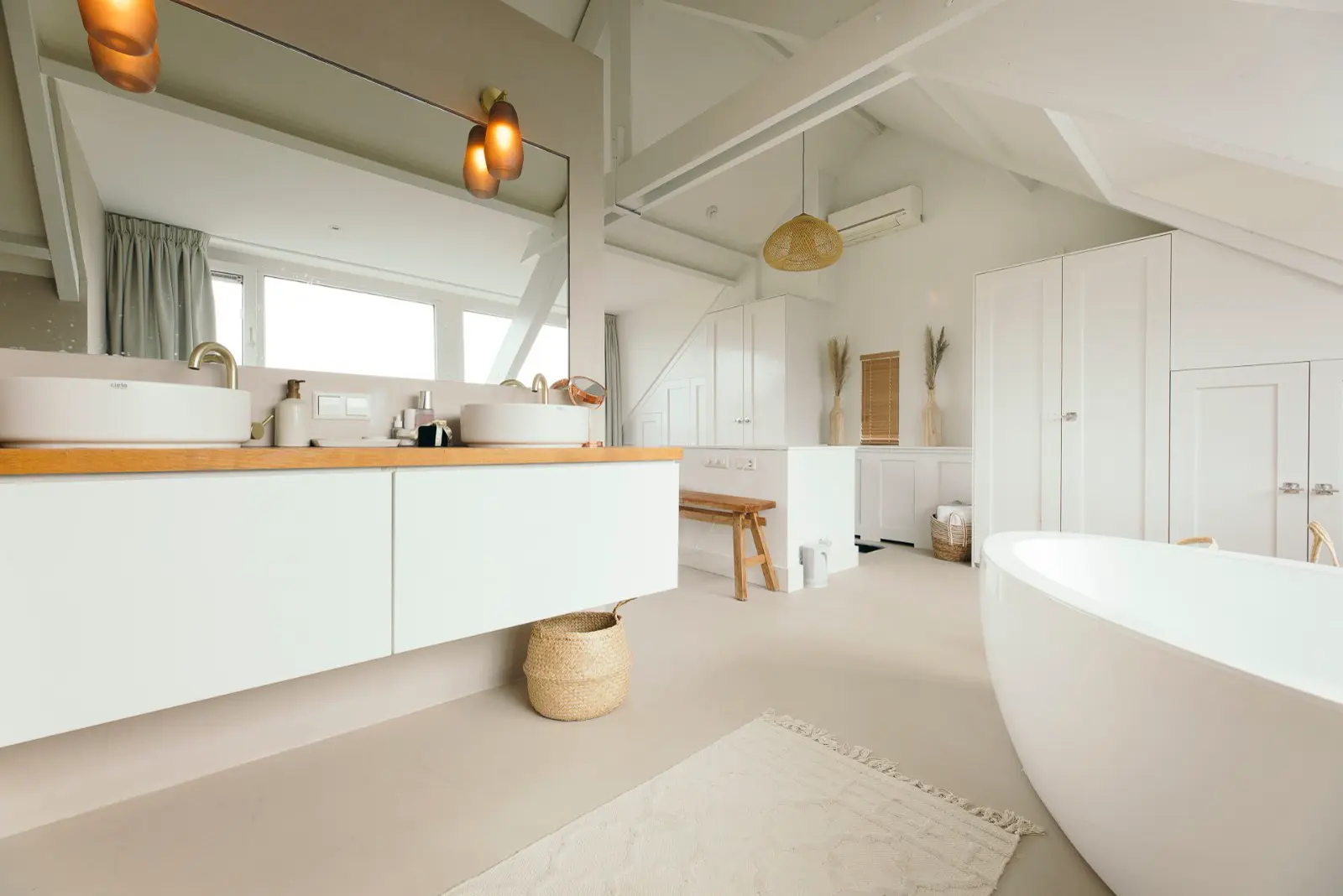 Ruime, licht inunderde badkamer met witte meubels, warme houten accenten en een doorlopende naadloze vloer in beige. De ruimte ademt minimalisme en rust uit met scherpe lijnen en perfecte ordening.