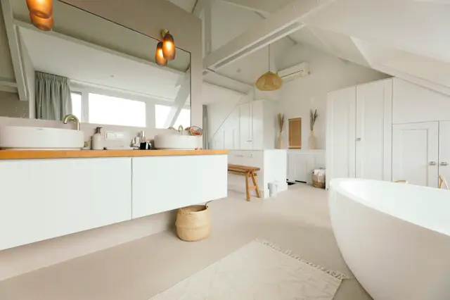 Ruime, licht inunderde badkamer met witte meubels, warme houten accenten en een doorlopende naadloze vloer in beige. De ruimte ademt minimalisme en rust uit met scherpe lijnen en perfecte ordening.