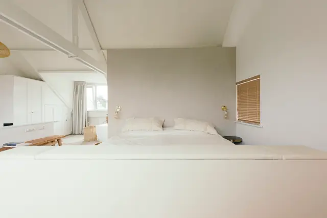 Serene slaapkamer met een grote naadloze vloer in warm wit, subtiel grijze wanden en natuurlijke lichtinval. Het ontwerp ademt rust uit met scherpe lijnen, gouden armaturen en minimale inrichting.