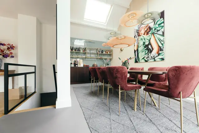 Ruime eetkamer met een strakke, lichte naadloze vloer in natuurtint. De ruimte ademt luxe en rust uit met minimalistisch design, grote raampartijen, bordeauxrode accenten en designer-elementen.
