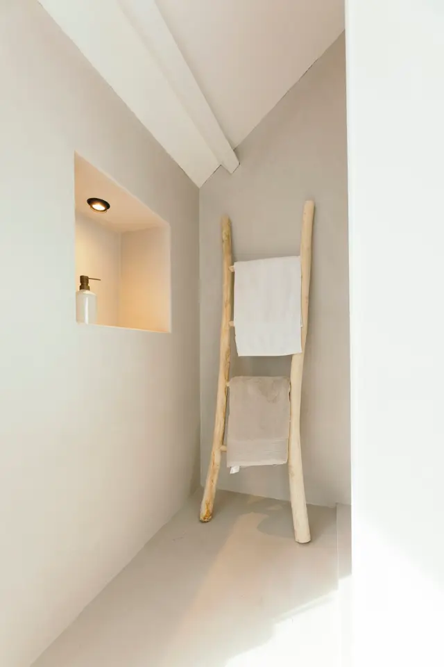 Een serene badkamerinrichting met zuivere witte wanden, naadloze vloer en een elegant houten handdoekrek. Warm licht uit de ingebouwde spiegel creëert een rustgevend en opgeruimd sfeer.