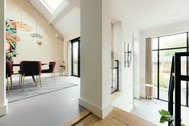Moderne, lichte woonruimte met naadloze vloer die van hout naar donkergrijs overgaat. Strakke architectuur met hoge ramen, natuurlijk licht en minimalistisch ingerichte zithoek met bordeauxrode stoelen.