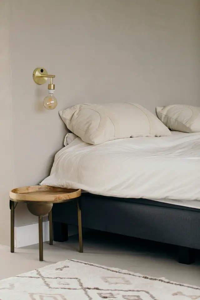 Een serene slaapkamer met naadloze lichte wand, strak opgemaakte bed in crème tinten en een houten bijzettafel. Een gouden wandlamp met bollamp geeft een subtiel accent.