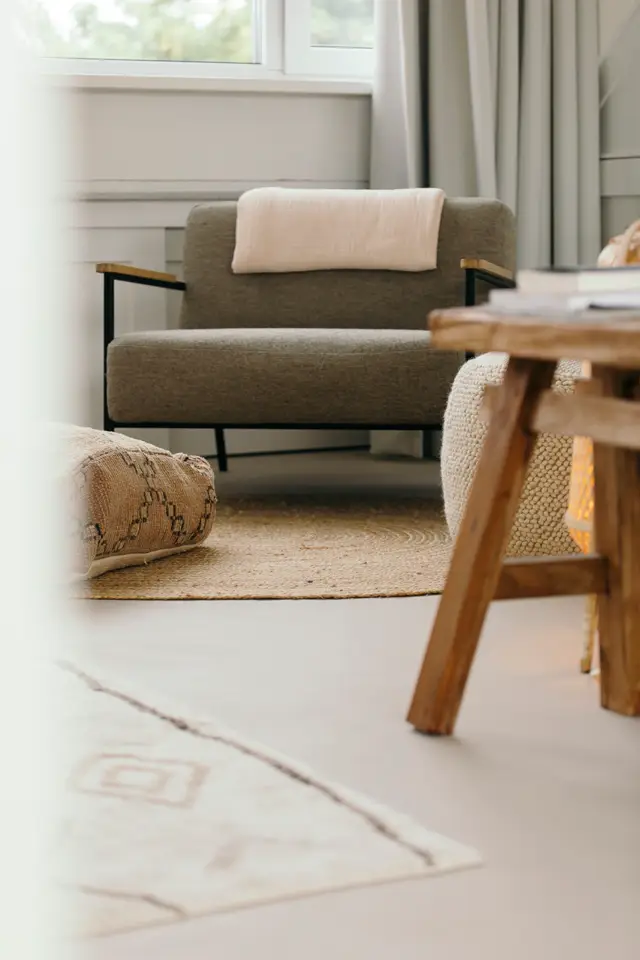 Serene woonkamer met minimalistische inrichting: een grijsgroene fauteuil, houten meubels en natuurlijke texturen op een lichte, naadloze vloer. Groot raam brengt overvloedig daglicht naar binnen.