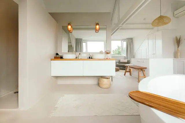 Ruime, licht badkamer met strakke naadloze wanden in warm wit. Dubbele wastafel met houten blad, grote spiegel en twee gouden wandlampen. Vrijstaand bad met houten rand, veel daglicht en minimalistisch ingericht.