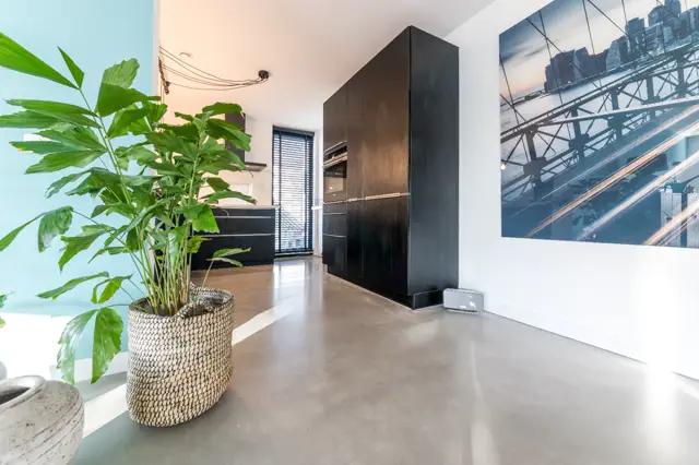 Strakke, lichte hal met glanzende naadloze vloer in warm grijs, zwarte designmeubels, groene plant in rieten pot en groot stadsfoto aan de wand. Alles perfect uitgelijnd en schoon.