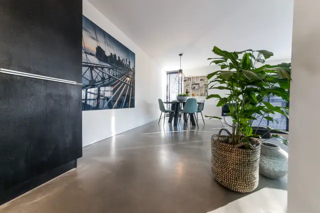 Ruime, licht ingevallen hal of woonzone met een grijstonige naadloze vloer. Links donkere wand met geïntegreerde details, rechts groene plant in naturel mand. Achtergrond toont eethoek en open kasten in warm witte tonen.