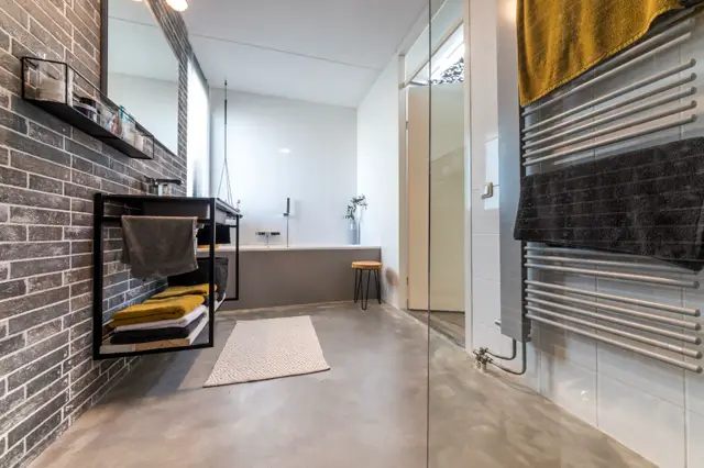 Ruime, modern ingerichte badkamer met grijs-bruine naadloze vloer en witte wanden. Strakke lijnvoering, designelementen in zwart en goud, volledige verlichting.