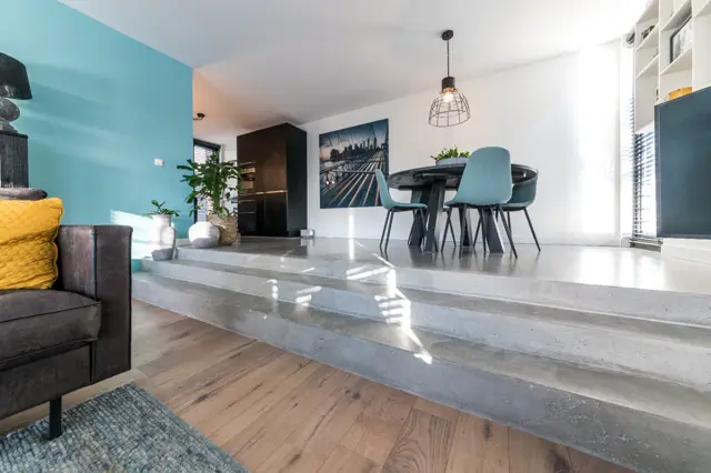 Strakke, lichte woonkeuken met naadloze vloer in grijs. Grote raampartijen zorgen voor helder daglicht, de ruimte oogt opgeruimd en minimalistisch met accenten in turquoise en zwart.