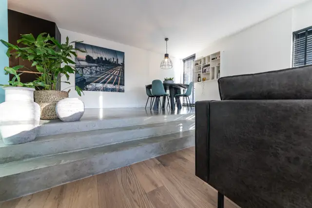 Strakke, lichte woonruimte met naadloze grijze wand die elegante ruwheid uitstraalt. Minimalistische inrichting met zwarte meubels, groene accenten en perfecte belichting.