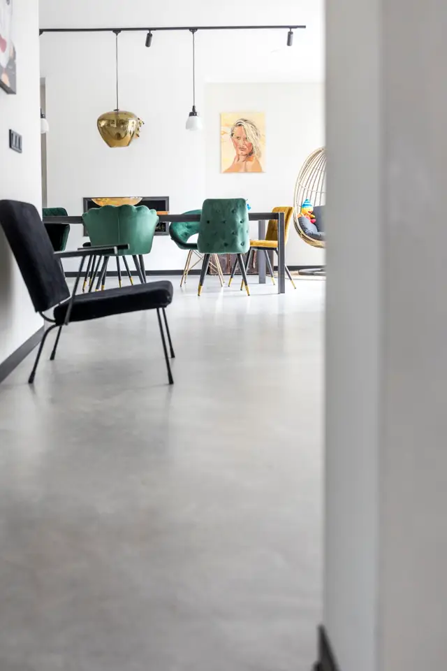 Ruime, licht en opgeruimd wooninterieur met naadloze vloer in licht grijs. Minimalistische inrichting met groen fluwelen stoelen, gouden hanglamp en scherpe lijnen.