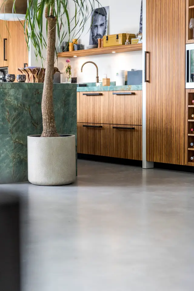 Een stijlvolle badkamer met een naadloze vloer in warm grijs, groengeaderde wand en houten accenten. Een witte plantenbak met minimalistisch design staat prominent in het ruimtelijke, lichte interieur.