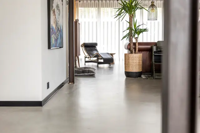 Een slanke doorgang met naadloze vloer in grijs, witte wanden en grote ramen die veel licht binnenlaten. In het achterliggende vertrek zijn designmeubels zichtbaar: een elegante loungestoel en verzorgde plantendecoratie.
