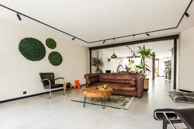 Ruime, open woonkamer met naadloze vloer in licht grijs. Strakke lijnen, minimalistische wanden en doordachte inrichting met vintage elementen en groene accenten creëren een serene, georganiseerde sfeer.