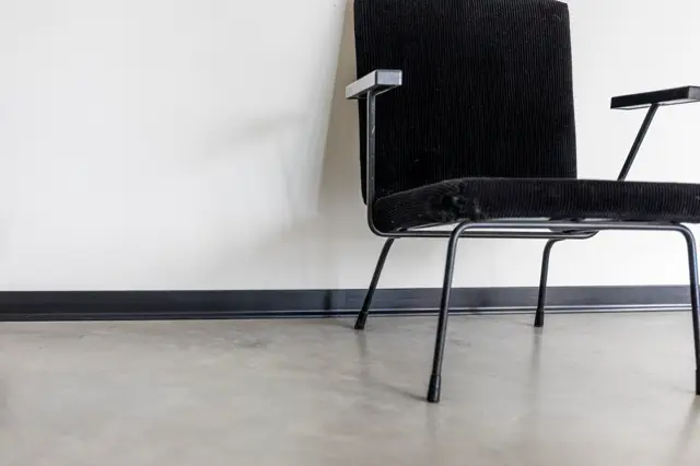 Een design-stoel met zwarte structuurstof en metalen frame staat op een lichte, vlekkeloze naadloze vloer tegen een witte wand. De compositie ademt rust en precisie uit.