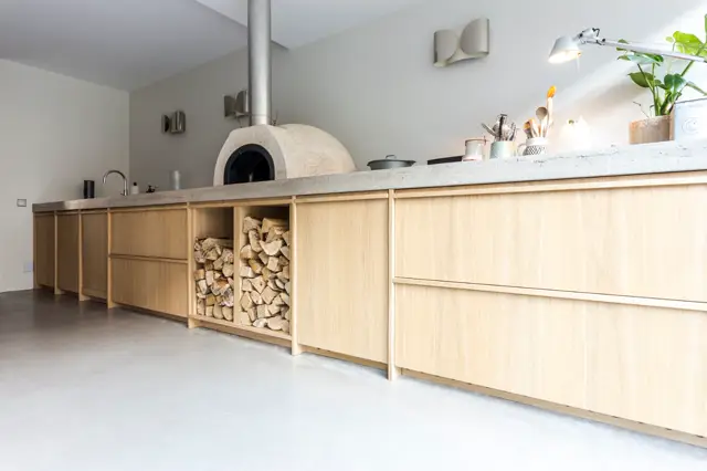 Strakke, lichte keuken met houten kasten, naadloze grijze vloer en natuurlijk licht. Een opvallende klassieke pizzaoven vormt het centerpiece, omgeven door opgeruimde werkbladen en verfijnde details.
