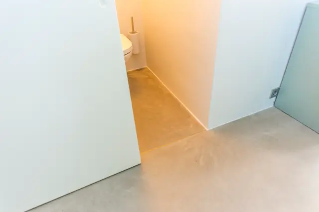 Een strak afgewerkte doorgang tussen twee ruimtes met naadloze vloeren in warme en grijze tinten. De witte wanden en scherpe lijnen creëren een minimalistisch, luxe interieur.