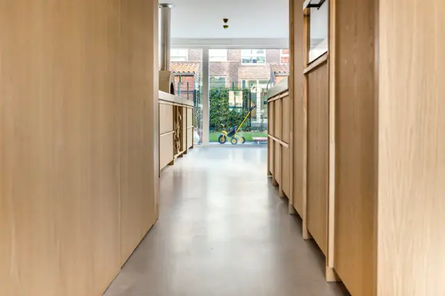 Een lichte, moderne gang met natuurlijk hout en een strakke naadloze vloer. Het perspectief loopt uit op een zonnig terras met groen en architectonische details.
