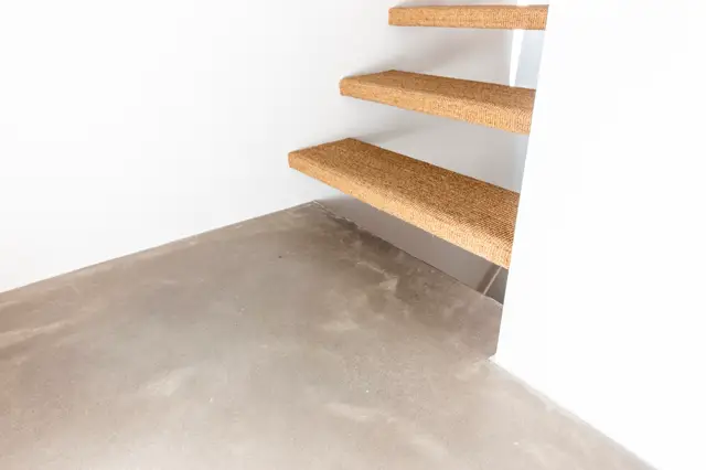 Minimalistische trapopstelling met natuurlijke jute treden op een strakke witte wand. De naadloze vloer in warm grijs loopt elegant door naar de trap, met perfecte lijnenvoering en subtiele schaduwwerking.
