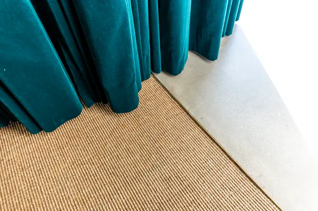 Een verfijnde ruimte met een strakke lichte naadloze wand, afgewerkt met een warm getextureerde vloer en opvallende tealkeurige gordijnen die elegant drapeëren. De scheiding tussen materialen is nauwkeurig uitgevoerd.