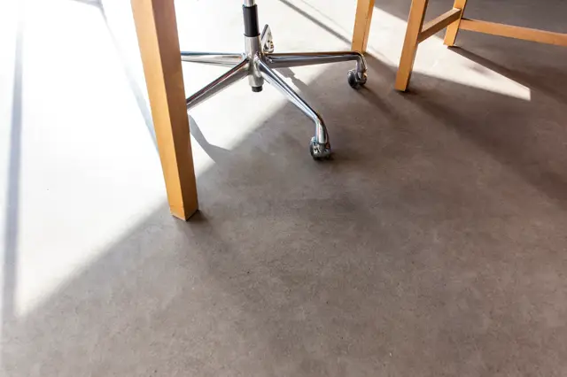 Een lichte, naadloze vloer met warme grijstint in een schone bedrijfsruimte. Natuurlijk licht valt in scherpe lijnen, houten meubelpoten en chromen stoelonderstel zorgen voor strak, modern contrast.