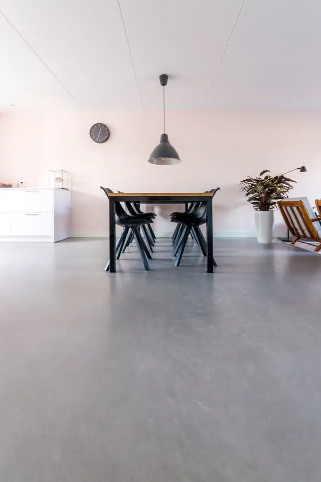 Lichte, open eetkamer met een strakke naadloze vloer in grijs, witte wanden en geometrisch plafond. Een zwarte eettafel met houten blad staat centraal, met minimalistisch ingericht interieur.
