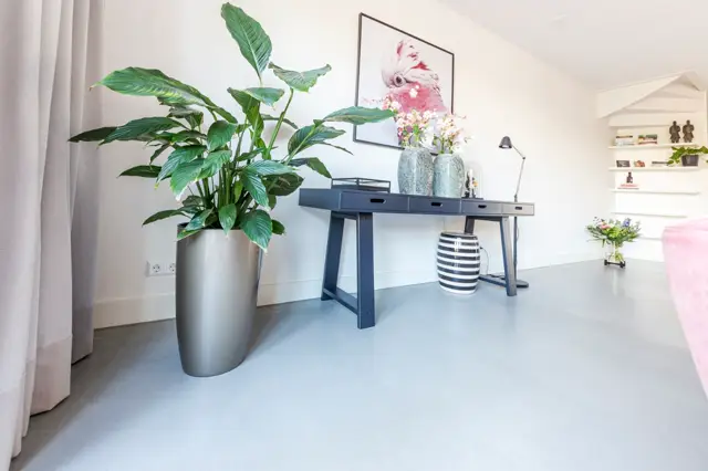 Licht, opgeruimd interieur met een naadloze lichte vloer die naadloos aansluit op witte wanden. Een strak design bureau met accessoires, groen plantwerk en subtiele decoratie creëren een rustige, luxe werksfeer.