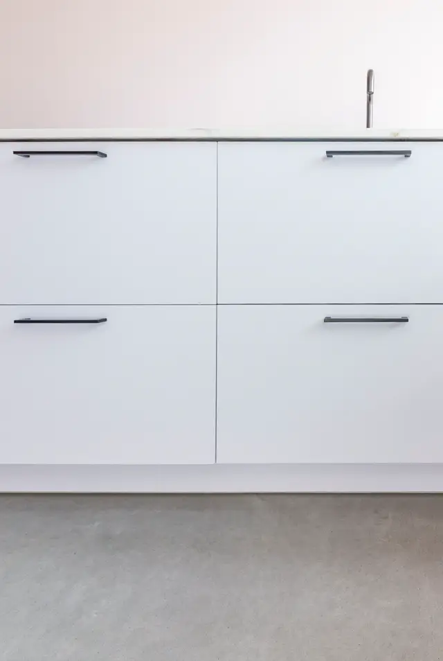 Strak witte keukenkasten met verfijnde metalen handgrepen en naadloze wand erboven in warm grijs. Naadloze vloer in betonlook vormt een rustgevend geheel.