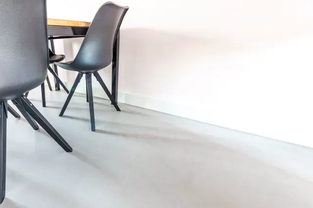 Strakke, lichte eetkamer met een naadloze vloer in warm grijs-beige en subtiele wand. Zwart-lederen stoelen rond een tafel met houten detail creëren een luxe, opgeruimde esthetiek.