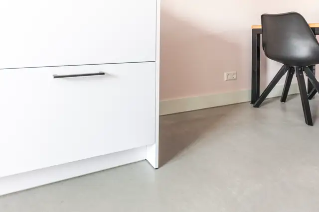 Een strak ingerichte hal met een naadloze lichte vloer die vrijwel onzichtbaar aansluit. Witte kasten, roze wand en modern zwart meubilair creëren een rustige, opgeruimde sfeer.