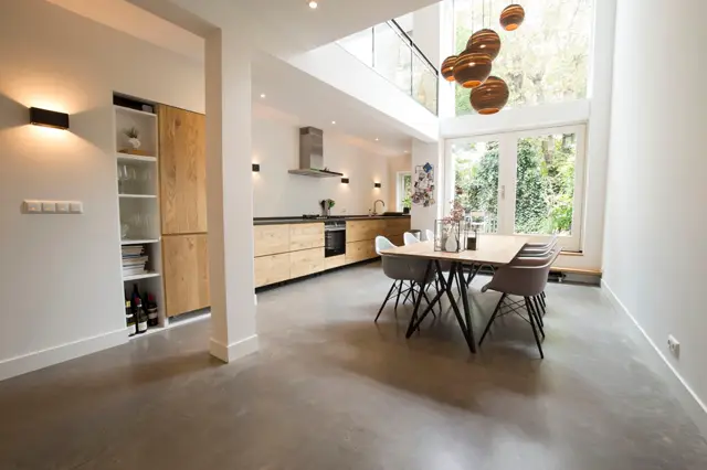 Ruime, lichtinundeerde open leefruimte met naadloze vloer, witte wanden en houten keukenfronten. Natuurlicht via grote raampartijen en skylight, strak design met warm houten accenten.
