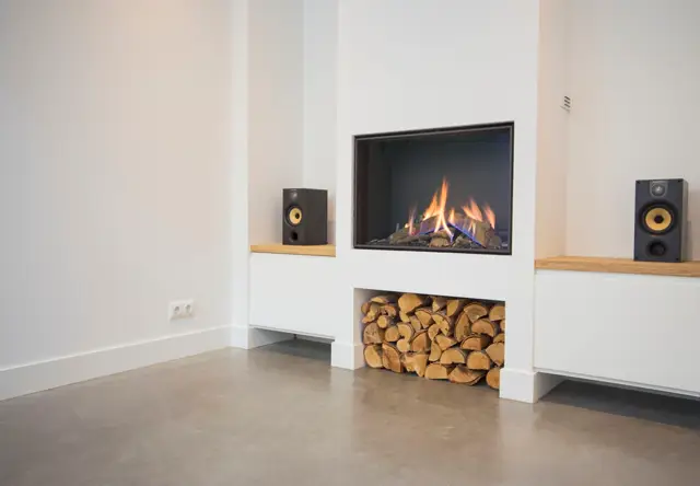 Strakke, lichte woonruimte met geïntegreerde haard, houten opberging voor brandhout en symmetrisch geplaatste speakers. De naadloze vloer en wanden creëren een rustgevend, exclusief interieur.