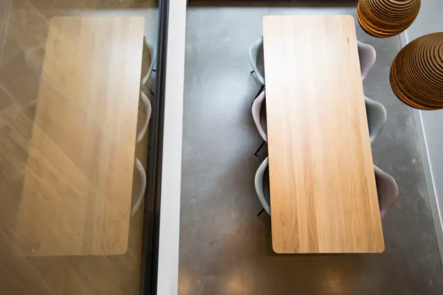 Luchtfoto van een strakke eetruimte met twee houten tafels op een naadloze grijze vloer, onderbroken door elegante witte stroken. Natuurlijk licht accentueert de schone lijnen en rustgevende sfeer.