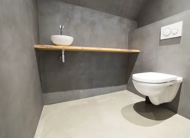 Strak ontworpen toiletruimte met naadloze grijze wanden en lichte vloer. Een wit ophangtarget boven een houten plank, aangevuld met minimalistisch design en warme materiaalcontrasten.