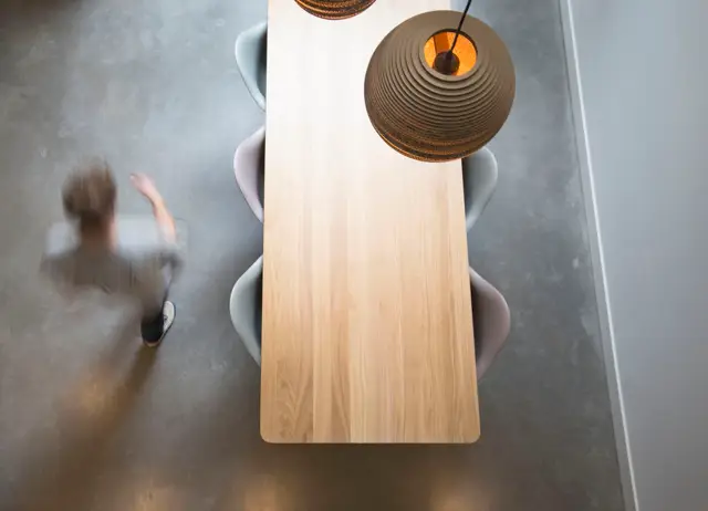 Vogelperspectief van een minimalistisch ingerichte eettafel met natuurlijke houten blad en designstoelen, geplaatst op een strakke grijze naadloze vloer. Hangende sculptuurlamp met warm licht voegt karakter toe.