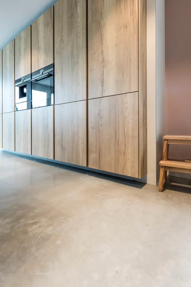 Strakke keukenfront in warm hout met ingebouwde oven, gecombineerd met een naadloze vloer in natuurlijke tint. Minimalistisch design met scherpe lijnen en elegante details.