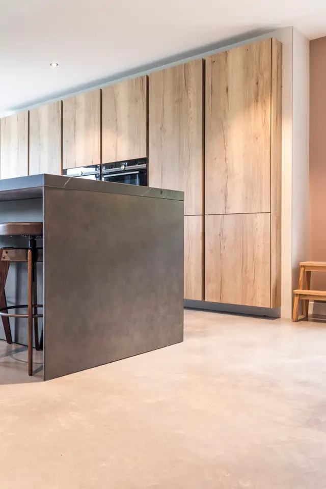 Moderne keukenruimte met een opvallende naadloze wand in warme houtstructuur en een grijze eiland. De vloer is naadloos afgewerkt, de lijnen zijn scherp en de belichting subtiel en professioneel.