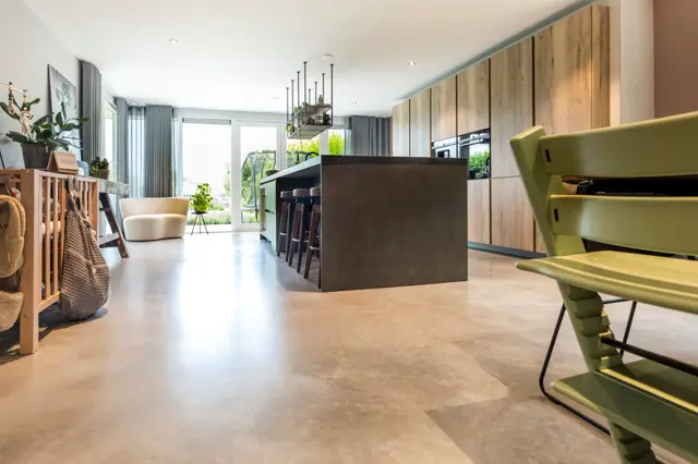 Ruime, open woon-keukenruimte met naadloze vloer in warme natuurtint. Strakke keukeneiland in donker contrast, luxe houten wandafwerkingen en grote raampartijen die zorgen voor heldere, diffuse daglichttoetreding.