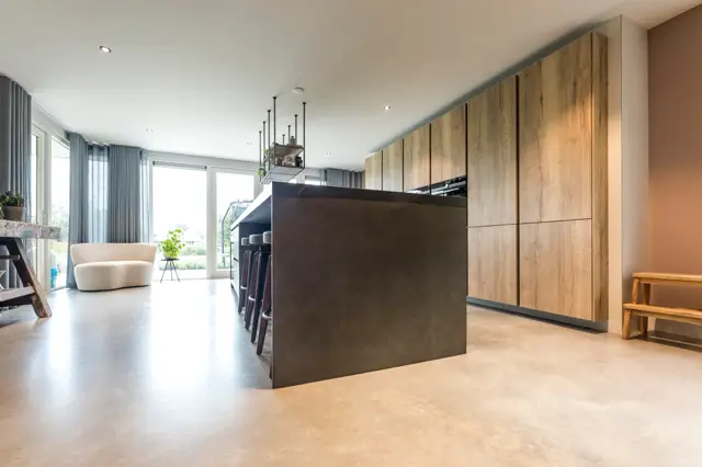 Lichte, naadloze vloer loopt seamless door een ruime, moderne open-plan woonkeuken met houten kastenwanden, donker eiland en groot raamoppervlak. De ruimte ademt rust en minimalisme.