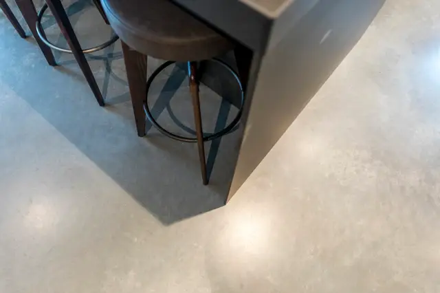 Minimalistische keukenhoek met een lichte naadloze vloer, elegant ontworpen met subtiele schaduwwerking. Moderne barstoel met zwart metalen frame creëert een strak, huiswerkaccent.