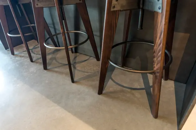 Luxe naadloze vloer in warm grijs met subtiele geometrische inlays, zichtbaar onder designer barstoelingen met donkerhouten poten en metalen voetsteun.