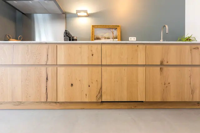 Elegante badkamerkast in natuurlijk hout met subtiele nerven en karakteristieke haarklinken, afgestemd op een rustieke grijze naadloze wandafwerking. Het geheel straalt sobere luxe en tijdloze vormgeving uit.