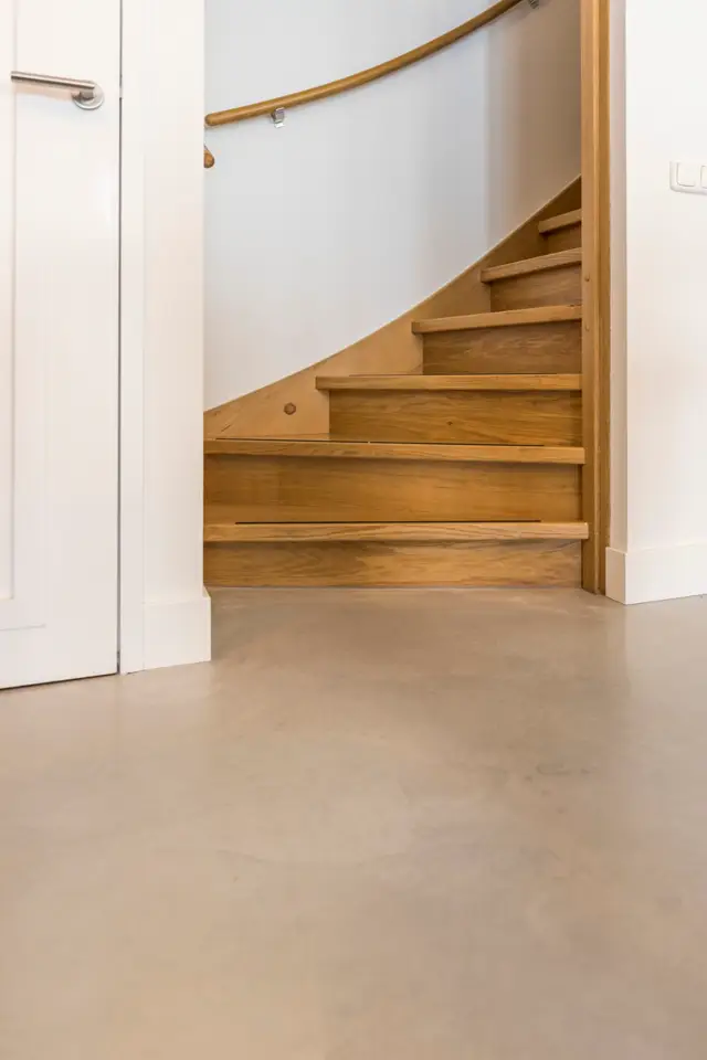 Een schone, lichte hal met houten trap en grijsbeige naadloze vloer. Wit stucwerk, houten leuning en minimalistische afwerking creëren een rustige, luxe sfeer.