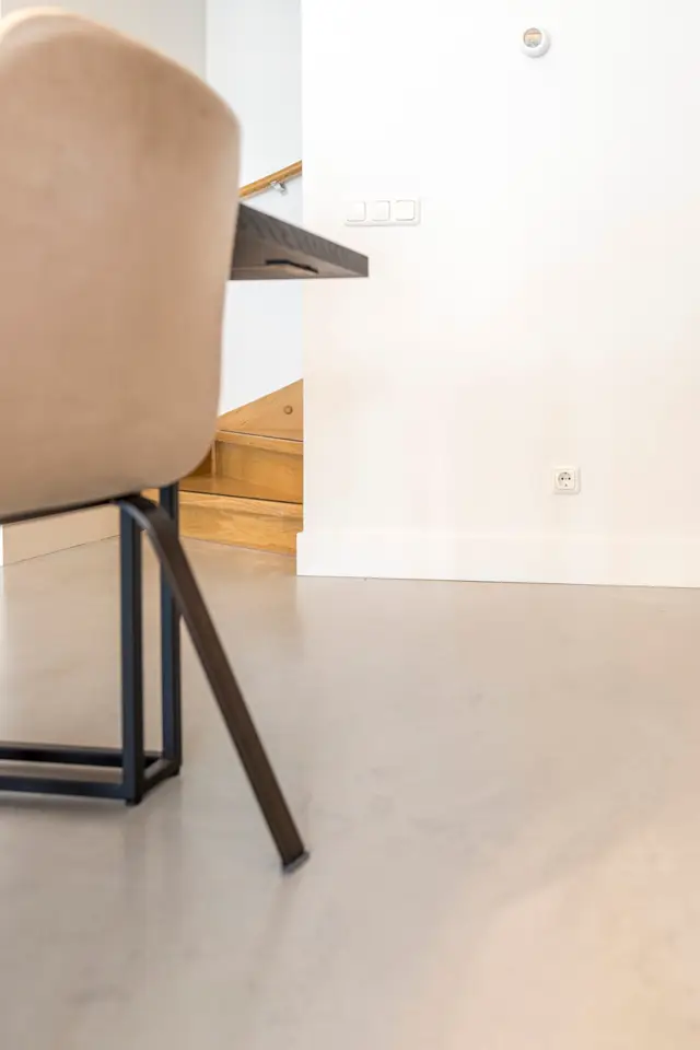 Een strak vormgegeven ruimte met een naadloze vloer in warm grijs, onderbroken door houten traptreden. Minimalistisch meubilair en witte wanden creëren een rustige, opgeruimde sfeer.