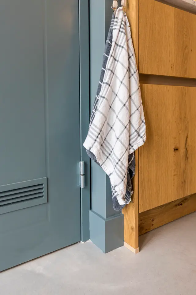 Detail van een strakke grijsgroene naadloze wand met minimalistisch deurkozijn in warm hout. De scherpe lijnen en rustieke houten accenten creëren een geraffeerd contrast.