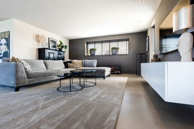 Ruime, licht ingerichte woonkamer met naadloze vloer in warme grijstinten. De ruimte ademt elegantie en minimalisme uit met strakke lijnen, perfect onderhouden meubels en professionele belichting.